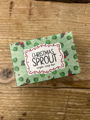 A Christmas Sprout vegan soap bar, featuring green sprout and holly designs on its packaging, is placed on a wooden surface.