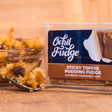 A pack of Sticky Toffee Pudding Fudge sits beside a bowl of sticky toffee pudding, with a fork holding a bite of the homemade fudge dessert in the foreground.
