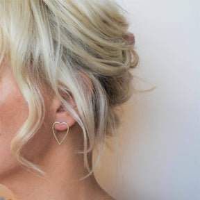 Close-up of a person with blonde hair in a loose updo, wearing MUKA Silver Heart Earrings. The large, delicate heart-shaped earrings stand out against the plain light background, with only the left side of the face and neck visible.