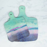 Two Arran Mini-Chopping Boards with handles, featuring a pastel landscape of mountains, water, and a cloudy sky in teal, purple, and pink tones, rest on a white marble surface.