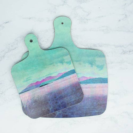 Two Arran Mini-Chopping Boards with handles, featuring a pastel landscape of mountains, water, and a cloudy sky in teal, purple, and pink tones, rest on a white marble surface.