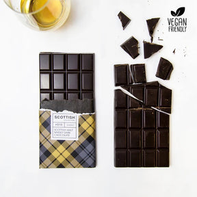 A whole handmade Dram of Whisky Dark Chocolate bar in a yellow tartan wrapper sits next to a broken bar and a glass of whisky; "Vegan Friendly" appears in the top right corner.