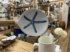 An Enamelled Wooden Spoon with a starfish design is held above a pitcher filled with white flowers, set against shelves displaying assorted home decor items in a store.