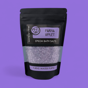 A black resealable pouch labeled "Parma Violet Epsom Bath Salts" with a purple design displays the salts inside. "Turns Water Purple" is written on the pack. Features a relaxing parma violet scent—ideal for eco-friendly gifting. Solid purple background.