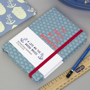 The Songs of the Mermaid Notebook is a blue A6 journal with a fish scale pattern, red elastic, and “THE SONGS OF THE MERMAIDS” in red. It’s displayed beside a ruler, pencil, anchor print pouch, and labeled “A LIFE ON THE OCEAN WAVES.”.