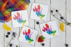 The Isle of Skye Watercolour Map Coaster, featuring a vibrant abstract design, sits on a white wooden surface with plaid cloth, blueberries, lemon wedges, a strawberry, and blue thistle—ideal for Scottish home decor.