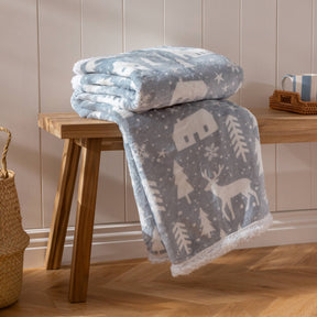 A folded Winter Walk Fleece Throw in Mist Blue (130 x 150 cm), featuring fluffy microfleece and white winter patterns of trees, reindeer, and snowflakes, rests on a wooden bench. Perfect for cozy Christmas décor, it is styled with a mug on a tray and a woven basket nearby on the wood floor.