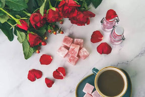 A marble table features red roses, rose petals, handmade Rose Turkish Delight cubes, two small bottles of pink liquid, and a green coffee cup with a matching saucer.