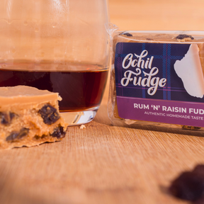 A glass of dark liquid sits next to a packaged block of Rum & Raisin Fudge and a partially eaten piece of butter fudge on a wooden surface. Raisins are visible in the rich, creamy Rum & Raisin Fudge.