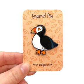 A hand displays a Puffin Enamel Pin attached to an orange card with a leaf pattern, reading "Enamel Pin" at the top and "neon-magpie.co.uk" at the bottom—showcasing this charming accessory.