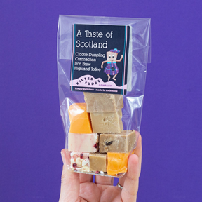 A hand holds a clear bag of Taste of Scotland Fudge, handmade in Scotland, featuring flavors like Clootie Dumpling, Cranachan, Iron Brew, and Highland Toffee against a purple background.