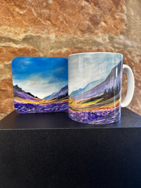 The Dawn of Hope Mug and matching coaster showcase a painted landscape of mountains, trees, purple wildflowers, and a cloudy sky on a textured brown stone background.