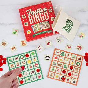 Overhead view of Festive Christmas Bingo, featuring a vibrant red box, green and orange bingo cards, red tokens, a cloth bag, and holiday-themed picture tiles. A hand places a red token on one of the cards—making Festive Christmas Bingo the perfect family game for your next holiday gathering.