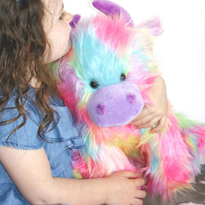 A young girl in a blue dress hugs and kisses the Highland Cow Soft Toy Large Rainbow - 30cm, featuring fluffy rainbow-colored fur and a purple face, nose, and horn, against a plain white background.