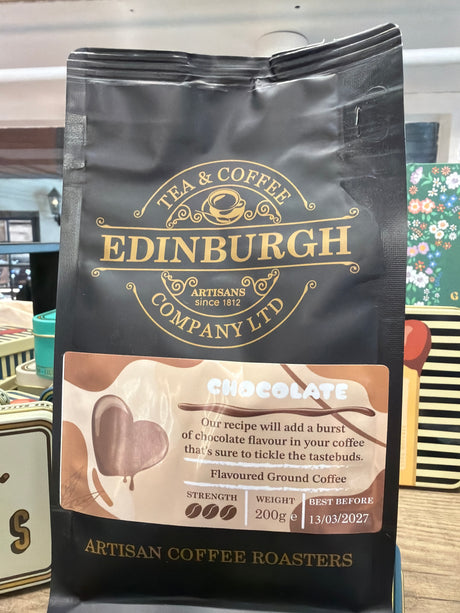 A black bag of Chocolate Flavoured Ground Coffee from Edinburgh Tea & Coffee Company Ltd, boasting a rich chocolate taste to delight your senses. The package displays strength, weight, and best before date, and is featured among other vibrant packages.