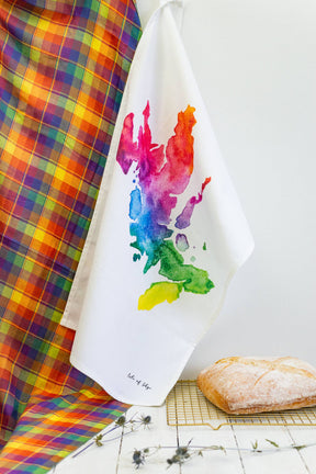 A Skye Watercolour Map Tea Towel with a vivid map design hangs beside a plaid cloth, while a loaf of bread cools nearby. “Isle of Skye” is inscribed at the bottom, making it ideal for any Scottish kitchen towel collection.
