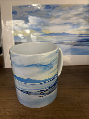 The Reflections of Arran Mug, featuring a seascape design, sits on a wooden surface in front of a matching print showing blue sky, clouds, distant mountains, and calm water.
