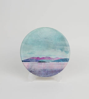 The Arran over Sound of Bute Ceramic Coaster Gift Boxed features a cork-backed, round design with a tranquil landscape of pink mountains, blue-green sky, and calm water, displayed on a clear stand against a white background.