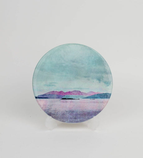 The Arran over Sound of Bute Ceramic Coaster Gift Boxed features a cork-backed, round design with a tranquil landscape of pink mountains, blue-green sky, and calm water, displayed on a clear stand against a white background.