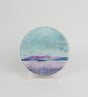 The Arran over Sound of Bute Ceramic Coaster Gift Boxed features a cork-backed, round design with a tranquil landscape of pink mountains, blue-green sky, and calm water, displayed on a clear stand against a white background.
