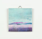 The Arran over Sound of Bute Gift Boxed ceramic art print features a serene lake, small islands, and distant purple mountains beneath a cloudy blue sky, beautifully presented in a ceramic tile and ready to display.