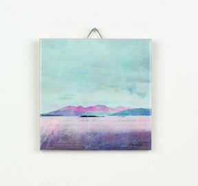 The Arran over Sound of Bute Gift Boxed ceramic art print features a serene lake, small islands, and distant purple mountains beneath a cloudy blue sky, beautifully presented in a ceramic tile and ready to display.