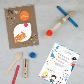 Make Your Own Pirate Catapult is an eco-friendly craft kit that includes wooden sticks, a clothespin, a spoon, a red pom-pom, a blue pom-pom, illustrated instructions, and comes packaged in a brown envelope featuring rocket and pirate-themed designs. Perfect for party favours and for developing fine motor skills in children, all components are displayed on a white surface.