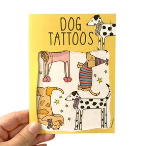 A hand holds a Dog Transfer Tattoos card featuring four cartoon dogs in playful outfits; these quirky dog transfer tattoos, perfect as party bag fillers, are visible through a window on the yellow card.