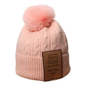 The Unisex Cable Bobble Hat: Pink is a light pink hat with a large fluffy pom-pom and a brown "Jess & Lou – The Cosy Hat" label, perfect for staying warm after wild swimming adventures.