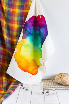 The Arran Watercolour Map Tea Towel, featuring a vivid rainbow stain, hangs beside a colorful plaid fabric while a loaf of bread cools on a rack below, all set against a crisp white background.