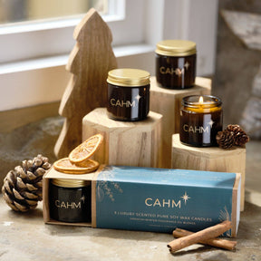 Christmas Candle Gift Set featuring three amber glass soy wax candles with gold lids on wooden blocks, with one candle lit and dried orange slices and cinnamon sticks in the foreground.