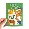 A hand holds a green packet labeled "Jungle Animal Transfer Tattoos," featuring quirky tattoos of a snake, tiger, toucan, orangutan, butterfly, and cheetah—making it a fun and unique gift option.
