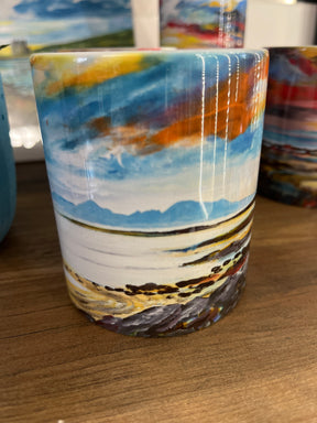The Towards the Warrior Mug is a ceramic mug featuring a painted landscape of a rocky shoreline, calm water, blue mountains in the distance, and a dramatic sky with orange, blue, and yellow clouds. Shown on a wooden surface.