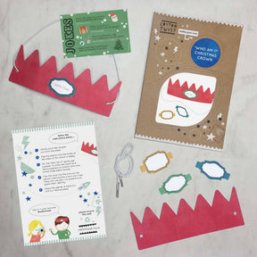 A flat lay of the Make Your Own ‘Who Am I?’ Christmas Cracker Crown kit, featuring red paper crowns, a fun ‘who am I?’ worksheet, an instruction sheet, a mini joke card, and a white elastic string on a marble surface—ideal for sustainable holiday activities or as an eco-friendly Christmas decoration.