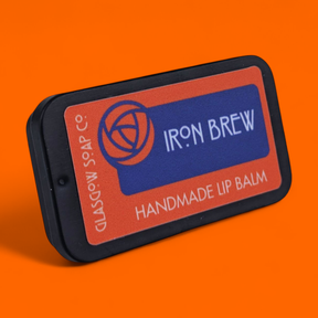 A rectangular tin of Iron Brew Lip Balm by Glasgow Soap Company rests on an orange background, featuring an orange and blue circular label.