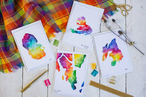 Four vibrant watercolor maps, featuring a Skye Map Greetings Card, are displayed on a white wooden surface among plaid fabric, scissors, a gold pen, ruler, binder clip, dried flowers, and watercolor pans.