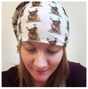 A person with light skin and straight, light brown hair wears the Highland Cow Multi-Functional Head & Neckwear. They have a nose piercing and are indoors against a plain background, looking down.