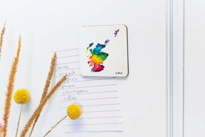 A Scotland Watercolour Map Magnet from Sarah Leask Studio holds a handwritten grocery list and dried yellow flowers on a white surface.