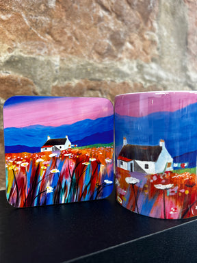 The Pink Sky Mug showcases white cottages, colorful wildflowers, and mountains under a vivid pink and blue sky, capturing the charm of a Scottish landscape with a stone wall backdrop.