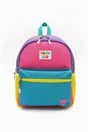 The Hairy Coo Kids Backpack has pink, purple, teal, and yellow panels with a blue front pocket, "HAIRY COO" label, and a small highland cow graphic in the lower right corner on a white background.