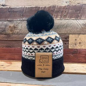 The Unisex Nordic Cosy Bobble Hat Black, featuring geometric patterns in black, grey, and tan and topped with a soft black pom-pom, rests on a wooden surface. A tag on the hat reads “Jess & Lou The Cosy Hat.” This unisex winter accessory blends warmth and Nordic-inspired style for chilly days.