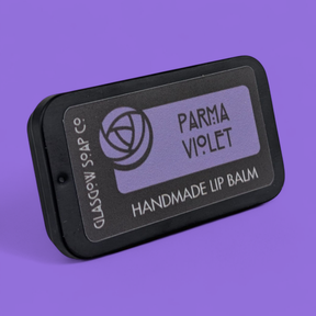 A rectangular tin of Parma Violet Lip Balm rests on a purple surface, featuring a circular rose logo and minimalist design. This natural lip balm is handcrafted by Glasgow Soap Co. and inspired by the Isle of Arran.