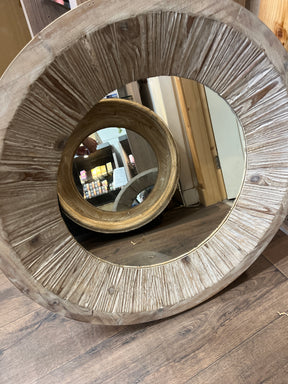 A large Round Wooden Mirror with a wooden frame sits on a wooden floor, reflecting a smaller round mirror to create a tunnel effect. Shelves with products and part of a doorframe are also visible in the background.