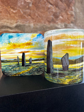 The Standing Stones Mug features matching artwork of standing stones in a grassy landscape beneath a dramatic sunset sky, with warm yellow and orange tones.