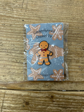 The Gingerbread Enamel Pin features a red bow tie and comes attached to a festive backing card with gingerbread cookies, all packaged in a clear wrapper and displayed on a wooden surface.