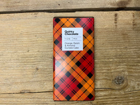 A Dundee Cake Chocolate with Orange, Raisin & Almond bar in orange, red, and black plaid packaging rests on a wooden surface, inspired by classic Dundee cake flavors.
