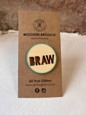 The Scottish Slang Wooden Brooch is a round, hand-painted wood accessory with "BRAW" in Scottish slang, presented on a kraft card labeled "WOODEN BROOCH, Hand-Painted." Includes glitterglitter.co.uk and social icons. A perfect Scottish touch!.