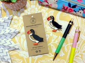 The Puffin Wooden Brooch — All That Glitters, presented on a labeled card and paired with a matching brooch, green and pink pen, patterned pouch, and decorative paper on a floral surface—an ideal thoughtful Scottish gift.