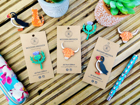 The Scottish Thistle Wooden Brooch — All That Glitters is hand-painted and displayed on a kraft card, surrounded by nature-inspired decor, reflecting the charm and heritage of Scotland.