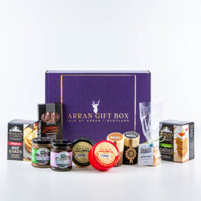 The Deluxe Favourites Arran Gift Box with gold lettering is displayed amidst Isle of Arran oatcakes, premium cheddar cheese, chutney, tablet, and preserves, all neatly arranged on a white background.